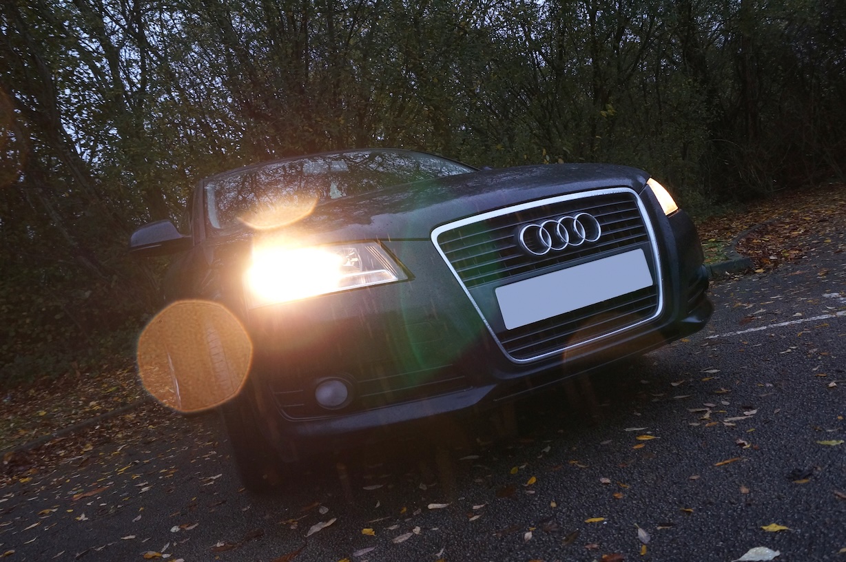 A photo of a black Audi A3 from the front in light rain. There are trees in the background and leaves on the floor. The light from the headlights, combined with water droplets on the lens produce some nice lens effects.