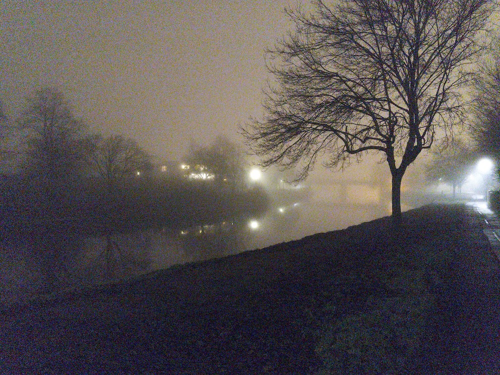 A shot taken from the side of a river on a foggy night. There is a bridge faintly visible through the fog in the distance.